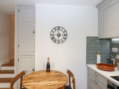A kitchen dining area with a round wooden table two chairs a bottle and two glasses a wall clock and kitchen cabinets at The Cottage on The Square in Chagford