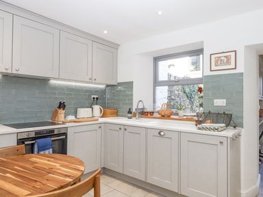 A kitchen with gray cabinets a wooden dining table a window over the sink and green tiled backsplash at The Cottage on The Square in Chagford