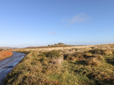 A winding road beside grassy fields with a rocky hill under a clear sky