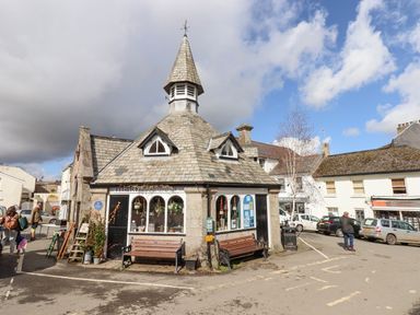 A stone building with a slate roof and a small tower in a town square with benches and parked cars at The Cottage on The Square in Chagford