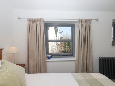 A bedroom window with beige curtains a bed with pillows and a blanket a bedside table with a lamp and books on the windowsill at The Cottage on The Square in Chagford