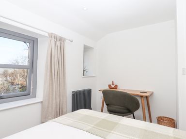 A bedroom with a bed, a wooden desk, a green chair, a window with beige curtains, and a radiator at The Cottage on The Square in Chagford