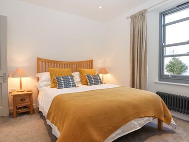 A bedroom with a wooden bed a yellow blanket pillows two bedside tables with lamps and a window with curtains at The Cottage on The Square in Chagford