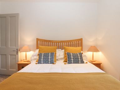 A bedroom with a wooden bed frame yellow and blue pillows white bedding lamps on wooden bedside tables and a closed door at The Cottage on The Square in Chagford