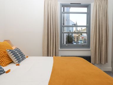 A bedroom with a bed with orange blanket and pillows next to a window with beige curtains at The Cottage on The Square in Chagford
