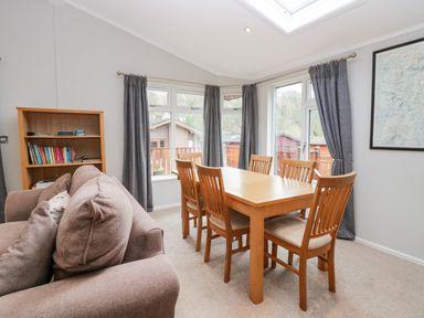 A dining area with a wooden table and six chairs next to windows with gray curtains and a bookshelf in a room at Highfield Lodge Kirkstone 30 Limefitt Park near Troutbeck