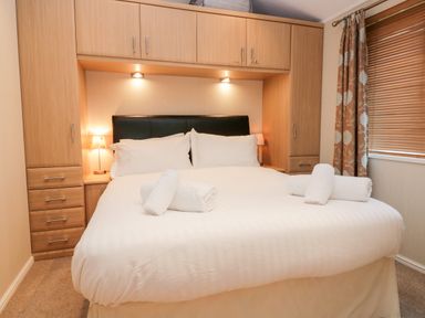 A bedroom with a double bed white linens rolled towels bedside lamps wooden cabinets and a window with blinds and floral curtains at Highfield Lodge Kirkstone 30 Limefitt Park near Troutbeck