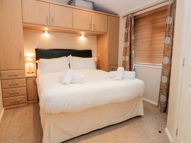 A bedroom with a double bed with white bedding and towels with built in cabinets and a window with brown blinds and floral curtains at Highfield Lodge Kirkstone 30 Limefitt Park near Troutbeck