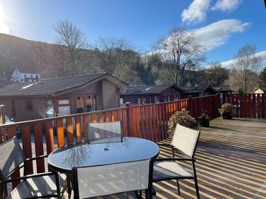 Outdoor deck with a glass table and chairs overlooking wooden cabins and trees at Highfield Lodge Kirkstone 30 Limefitt Park near Troutbeck