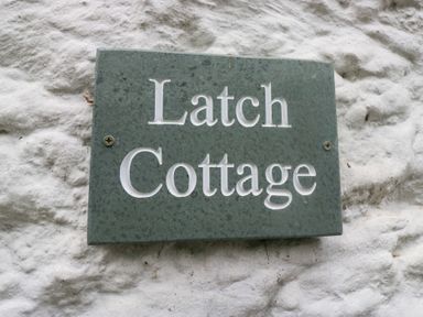 A green plaque with the words Latch Cottage on a white textured wall at Latch Cottage in Keswick