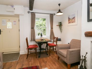 A small dining area with two wooden chairs with red cushions around a wooden table near a window with curtains and an armchair in a room at Latch Cottage in Keswick