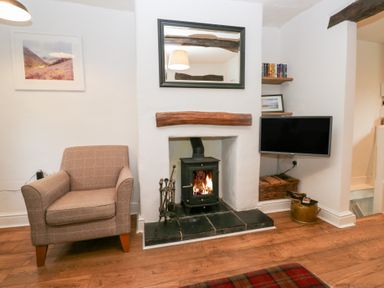 A living room with an armchair a wood-burning stove under a wooden mantel a wall-mounted television and wooden flooring at Latch Cottage in Keswick