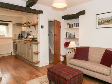 A kitchen and living room area with wooden beams a sofa with cushions a plaid ottoman and open shelves with books at Latch Cottage in Keswick