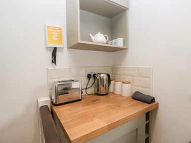 A kitchen counter with a toaster kettle canisters and a towel under a wall shelf with teapot and mugs at Latch Cottage in Keswick