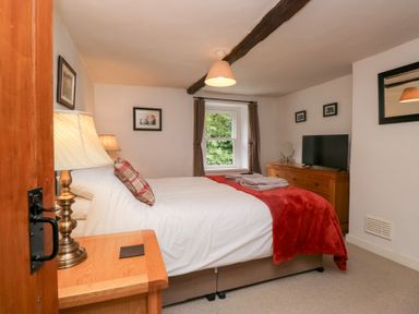 A bedroom with a bed with white and red bedding a wooden nightstand with a lamp a window and a wooden dresser with a television at Latch Cottage in Keswick