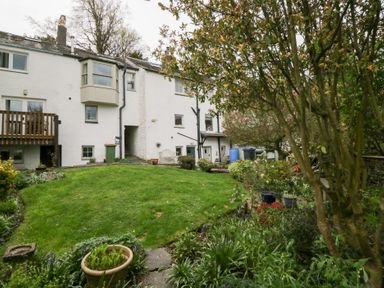 A garden with grass and plants in front of a white house with a wooden balcony and bay window at Latch Cottage in Keswick