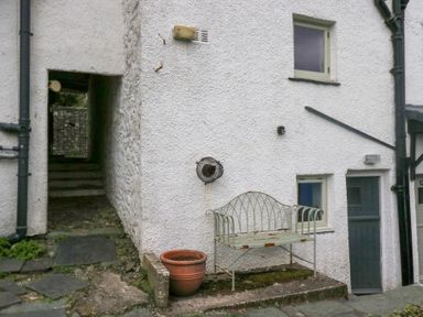 An outdoor area with a metal bench and a large empty flower pot by a white textured wall with a gray door and two windows at Latch Cottage in Keswick