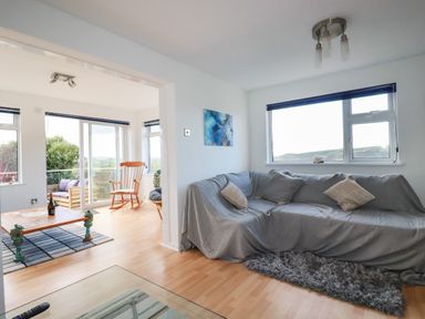 A living room with a gray covered sofa and pillows next to a window and an adjoining room with a rocking chair and coffee table at Seacroft in Crantock