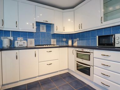 A kitchen with white cabinets blue tiled walls and floor a microwave kettle toaster and double oven at Seacroft in Crantock