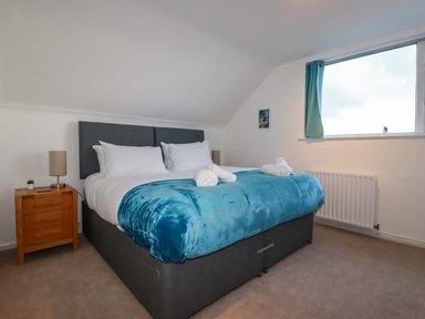 A bedroom with a double bed with blue blanket white pillows and rolled towels next to a wooden bedside table with a lamp and a window with blue curtains at Seacroft in Crantock