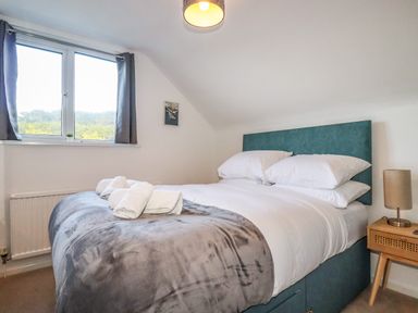 A bedroom with a bed with white pillows and gray blanket next to a wooden nightstand with a lamp and a window with gray curtains at Seacroft in Crantock