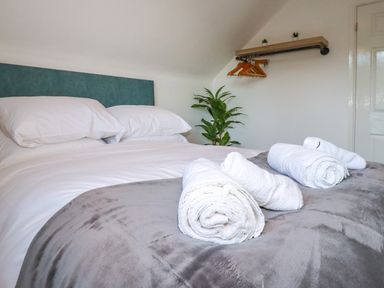 A bedroom with a bed with white pillows and sheets grey blanket and rolled white towels on the bed a plant near the wall and a wooden shelf with hangers on the wall at Seacroft in Crantock