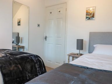 A bedroom with two beds a wooden nightstand with a lamp a door a wall mirror and artwork at Seacroft Crantock