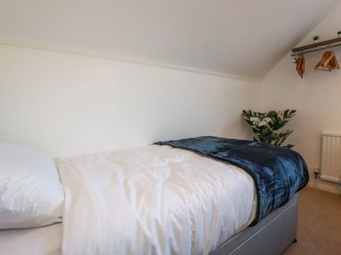 A bedroom with a bed covered with white bedding and a dark blue blanket with a plant and hanging clothes in the corner at Seacroft in Crantock