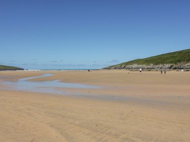A sandy beach with a small stream and people walking near grassy cliffs at Seacroft in Crantock