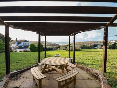 A wooden picnic table with benches under a wooden pergola on a patio overlooking a grassy garden with houses and hills in the distance at Seacroft in Crantock