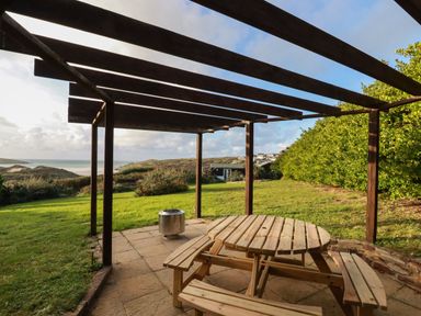 A wooden pergola with a round wooden picnic table and benches on a patio overlooking grass and hills at Seacroft in Crantock