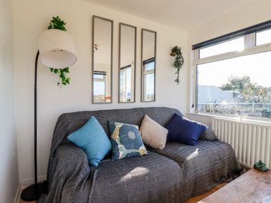 A living room with a sofa covered with a dark fabric and cushions by a window with a lamp and three vertical mirrors on the wall at Seacroft in Crantock