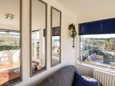 A room with a sofa covered by a throw and a blue pillow near a window with blue blinds and three vertical mirrors on the wall at Seacroft in Crantock