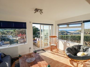 A sunlit living room with wooden rocking chair papasan chair wooden table and large windows overlooking outdoor seating and landscape at Seacroft in Crantock
