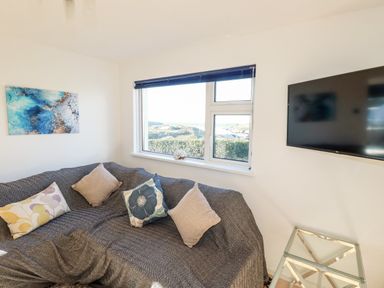 A living room with a sofa covered by a gray throw and decorated with pillows near a window and a wall-mounted TV at Seacroft in Crantock