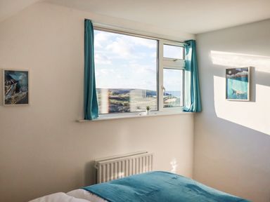 A bedroom with a bed covered in teal bedding next to a window with teal curtains and two framed pictures on the walls at Seacroft in Crantock