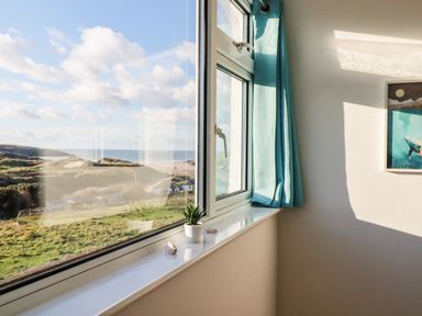 A window with blue curtains overlooking a grassy landscape and sea with a small plant on the windowsill at Seacroft in Crantock