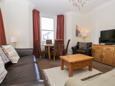 A living room with brown leather sofa and armchair wooden coffee table dining table with four chairs and television on wooden cabinet at Carlton House Apartment 1 in Keswick