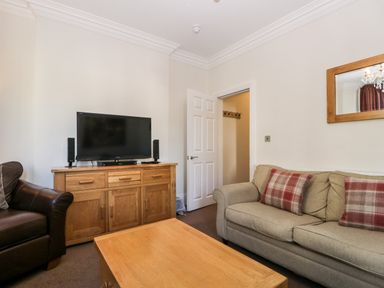 A living room with a gray sofa with plaid pillows a wooden coffee table a wooden cabinet with a TV and speakers and an open door at Carlton House Apartment 1 in Keswick
