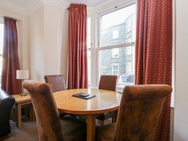 A dining area with a round wooden table and four chairs near windows with patterned red curtains at Carlton House Apartment 1 in Keswick