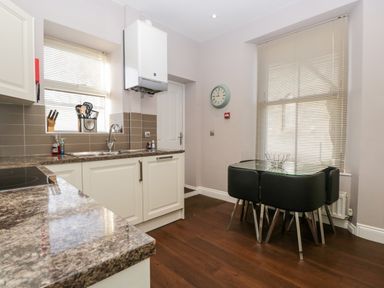 A kitchen with a marble countertop white cabinets a glass dining table with black chairs and a wall clock at Carlton House Apartment 1 in Keswick