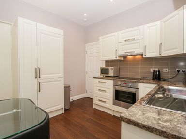 A kitchen with white cabinets a microwave an oven a black kettle a stainless steel sink and a glass table at Carlton House Apartment 1 in Keswick