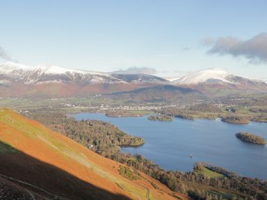 A lake surrounded by forest and hills with snow-capped mountains in the background at Carlton House Apartment 1 in Keswick