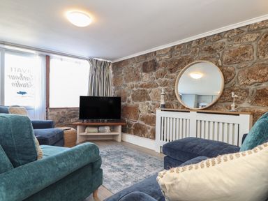 A living room with a stone wall a round mirror blue armchairs a television and striped curtains at Harbour Studio in Sennen Cove
