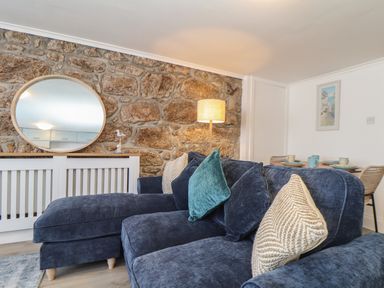A living room with a blue sectional sofa stone wall a round mirror a floor lamp and a dining table at Harbour Studio in Sennen Cove