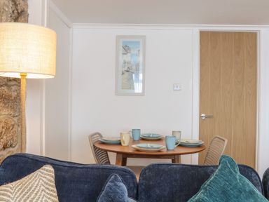 A dining area with a wooden round table set with four plates and mugs surrounded by woven chairs and a blue sofa in the foreground at Harbour Studio in Sennen Cove