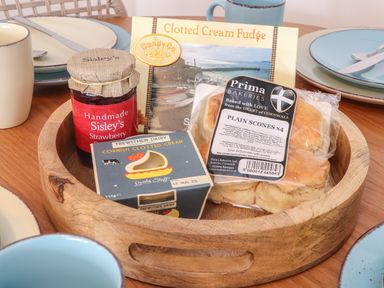 A wooden tray with strawberry jam, clotted cream, plain scones, and clotted cream fudge on a wooden table with plates and mugs at Harbour Studio in Sennen Cove