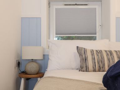 A bed with white pillows and a striped cushion next to a small wooden bedside table with a round lamp and a window with a blind behind the bed at Harbour Studio in Sennen Cove