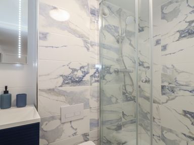 A bathroom with a shower enclosed by glass walls a showerhead mounted on marble-patterned tiled walls and a small sink with blue soap dispenser and cup