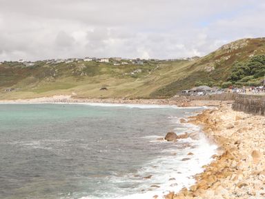 A rocky coastline with waves hitting the shore and a sandy beach with houses on a hill at Harbour Studio in Sennen Cove